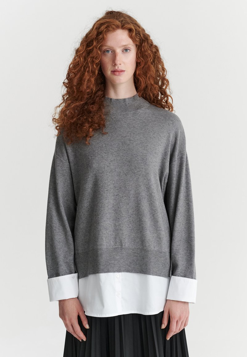 Femme aux longs cheveux roux bouclés portant un pull gris superposé à une chemise blanche et une jupe plissée noire, debout devant un fond uni.