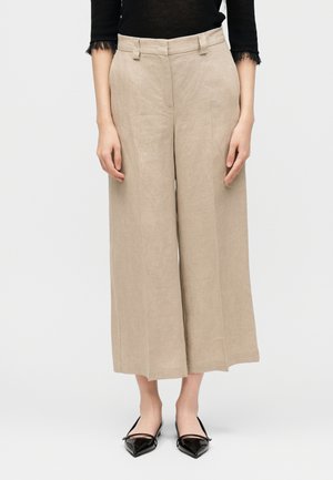 Pantalon beige large cropé avec passants, porté avec des ballerines noires à bout pointu et un haut noir avec manches en dentelle trois-quarts.