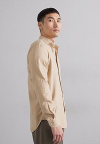 Beige långärmad skjorta med knappar och klassisk krage, slät yta och bröstficka, visad mot en neutral bakgrund.