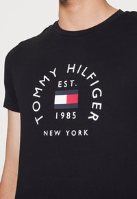Camiseta de algodón negra con un logo blanco circular que presenta "TOMMY HILFIGER", "EST. 1985" y un diseño rectangular de bandera en rojo, blanco y azul.