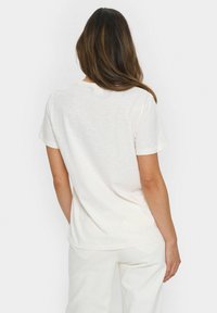 Mujer con cabello largo castaño, vistiendo una camiseta blanca de manga corta simple y pantalones blancos, de espaldas contra un fondo blanco.