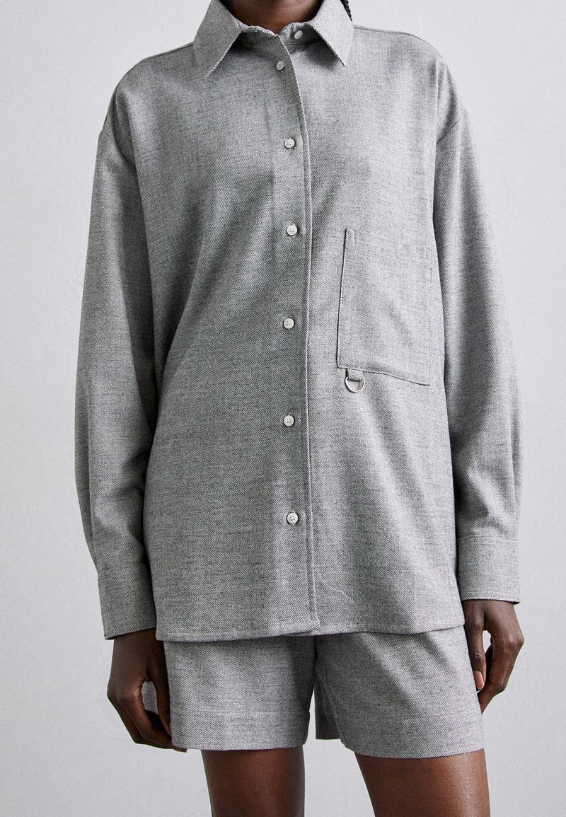 Chemise grise à boutons avec une poche poitrine mettant en avant un détail en anneau métallique, assortie de shorts gris fabriqués dans un tissu texturé.