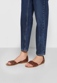 Sandales en cuir tressé marron avec bout ouvert et design sans talon, associées à un jean en denim bleu. Les sandales ont une semelle plate.