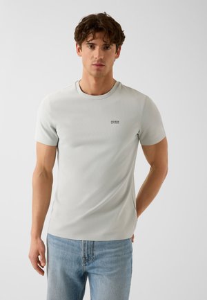 Jeune homme aux cheveux bruns portant un t-shirt gris clair à manches courtes et un jean bleu, debout devant un fond blanc uni.