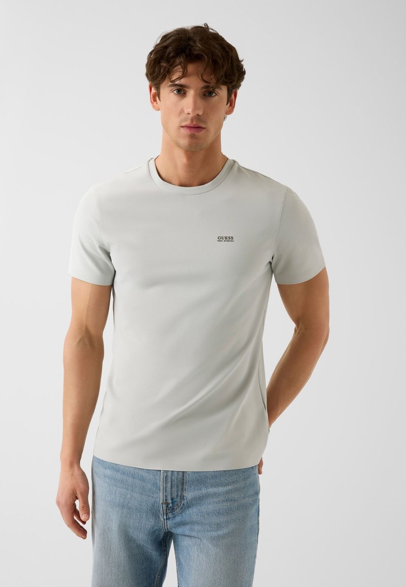Jeune homme aux cheveux bruns portant un t-shirt gris clair à manches courtes et un jean bleu, debout devant un fond blanc uni.