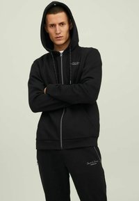 Schwarze Zip-Hoodie mit einer Kordelzugkapuze, grauer Logo-Stickerei und gerippten Bündchen. Dazu passende schwarze Jogginghosen. Glatte Stoffbeschaffenheit.
