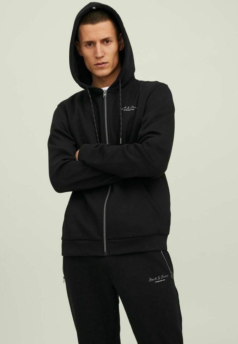 Schwarze Zip-Hoodie mit einer Kordelzugkapuze, grauer Logo-Stickerei und gerippten Bündchen. Dazu passende schwarze Jogginghosen. Glatte Stoffbeschaffenheit.