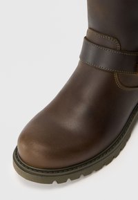 Bota de tornozelo em couro castanho com biqueira arredondada, superfície texturizada e sola de borracha resistente. Apresenta um detalhe em tira com costura em contraste.