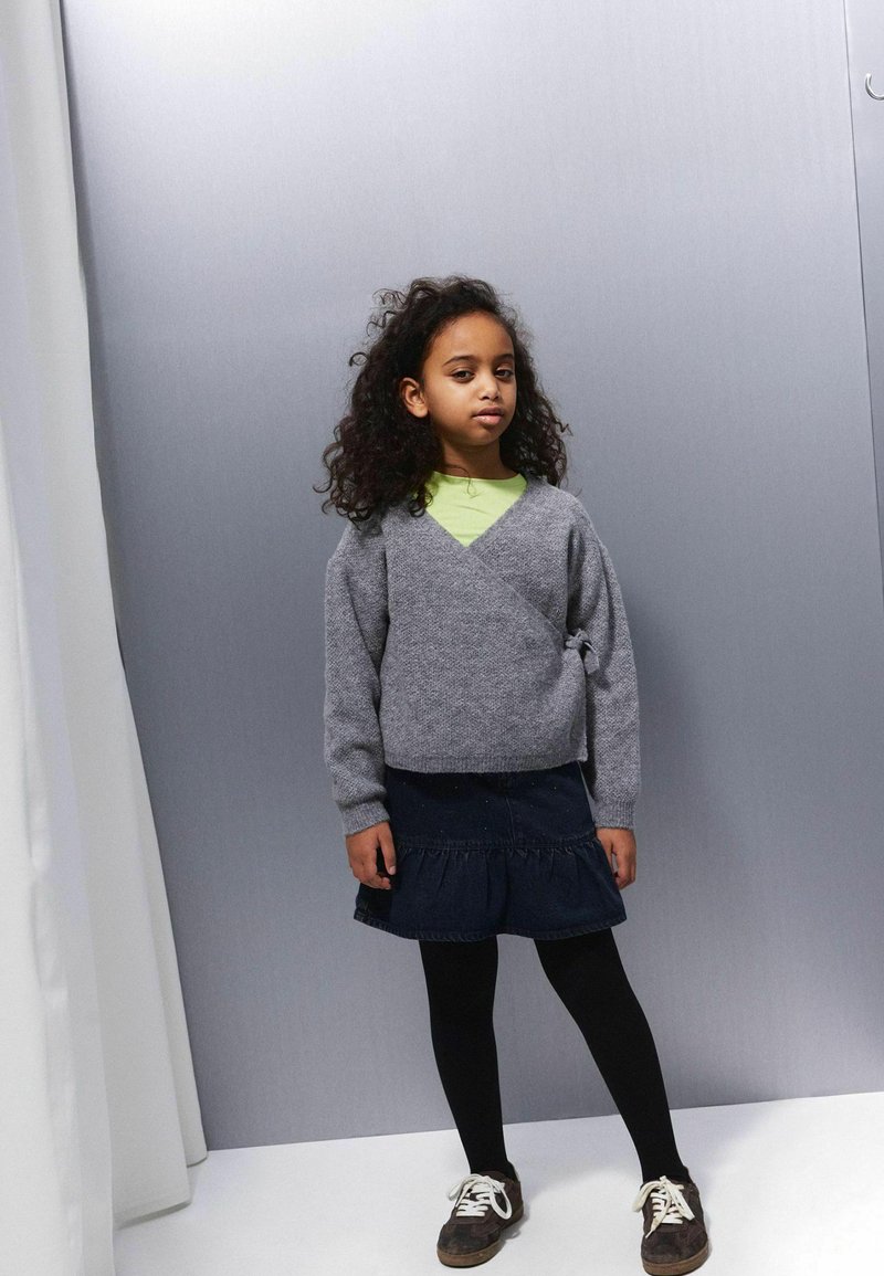 Mala djevojčica s kovrčavom kosom nosi sivi džemper, tamnoplavu traper suknju, crne tajice i smeđe tenisice, a stoji protiv sive pozadine.