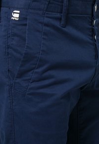 Tmavě modré kalhoty s hladkou texturou a jemným leskem, s logem na přední straně a standardním zapínáním na knoflíky.