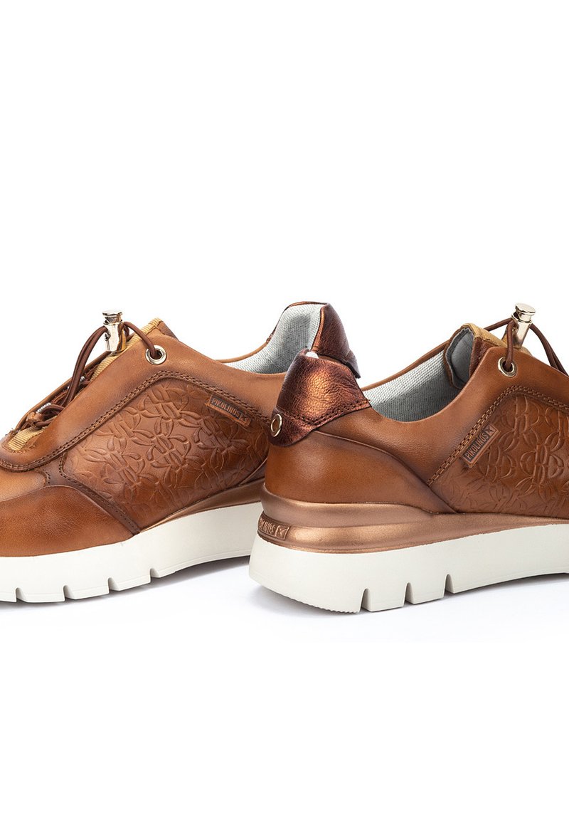 Zapatillas de cuero marrón con un patrón floral texturizado, acentos metálicos y suelas de goma blanca. Presentan un diseño de cordones y un collar acolchado.