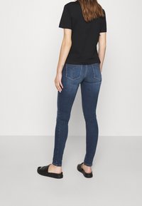 Camiseta negra, jeans de denim azul ajustados con bolsillos traseros y sandalias negras de deslizar. Tejido suave con un lavado oscuro y ligero estiramiento.