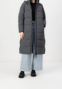 Gewatteerde lange grijze jas met een capuchon, voorzien van een rits aan de voorkant, zijzakken en elastische boorden, gecombineerd met lichtblauwe jeans met wijde pijpen en zwarte schoenen.