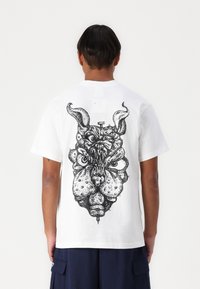 White cotton t-shirt featuring a large black graphic of a creature's face with horns and exaggerated facial features on the back.