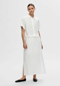 Robe en lin blanc à manches courtes, devant à boutons, taille à cordon, avec fente sur le côté, assortie à des chaussures plates noires.