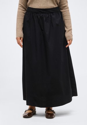 Femme portant une longue jupe noire, un pull beige et des chaussures plates marron avec brides à la cheville, se tenant sur un fond blanc uni.