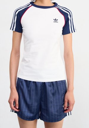 Osoba nosząca białą koszulkę Adidas z granatowymi rękawami i paskami, w połączeniu z granatowymi spodenkami w cienkie paski z bocznymi paskami.