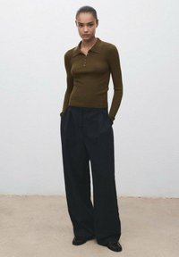 Brązowy sweter w paski z kołnierzykiem, zestawiony z czarnymi spodniami o wysokim stanie i szerokiej nogawce. Modelka stoi z rękami w kieszeniach na tle o fakturze.