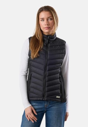 Jonge vrouw met lang haar, gekleed in een zwart doorgestikt vest over een wit overhemd met lange mouwen en een blauwe spijkerbroek, staande tegen een effen achtergrond.