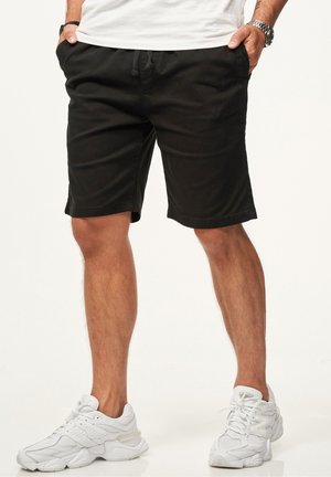 Mann trägt schwarze Shorts mit Kordelzug, weiße Sneaker und ein weißes Hemd, steht mit den Händen in den Taschen vor weißem Hintergrund.