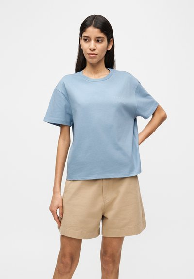 Ανοιχτό μπλε βαμβακερό μπλουζάκι με στρογγυλή λαιμόκοψη και κοντά μανίκια, σε συνδυασμό με καφέ σορτς. Απλός σχεδιασμός με λιτό κείμενο.