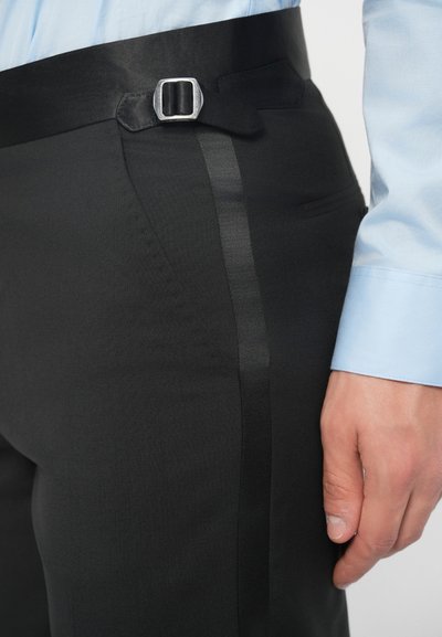 Detailní záběr na osobu v černých šitých kalhotách se stříbrnou sponou na boku, přičemž je vidět rukáv světle modré košile.