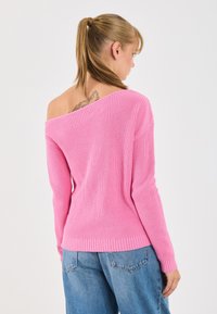Giovane donna con coda di cavallo bionda, che indossa un maglione rosa off-shoulder e jeans blu, mostrando un tatuaggio sulla parte superiore della schiena contro uno sfondo bianco.