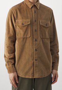 Brun kordfløjls skjorte med knaplukket krave, to forlommer og mørke knapper, med lodrette striber i stofteksten.