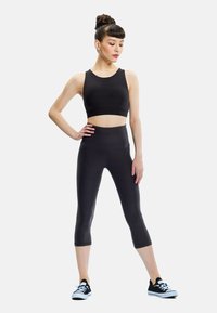 Woman in black cropped tank top, high-waisted capri leggings, and black sneakers standing with hand on hip looking down.