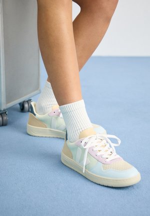 Persona con zapatillas de colores pastel y calcetines blancos de pie sobre una alfombra azul cerca de un objeto metálico con ruedas.