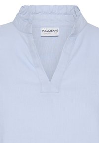 Blouse bleu clair avec un col volanté et un décolleté en V, avec une étiquette de la marque Pulz Jeans à l'intérieur du col.