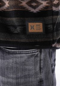 Fleece-pullover med ett mönstrat design i mörka toner och en brun läderlapp, parat med grå denimjeans.