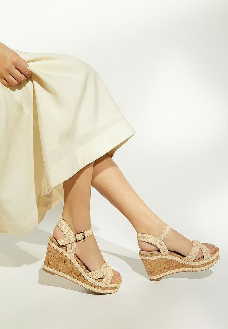 Sandales compensées beiges avec une semelle en liège, dotées de brides tressées et d'une fermeture à boucle. Le matériau supérieur semble texturé et tissé.