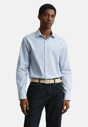 Jeune homme portant une chemise à rayures verticales bleues et blanches rentrée dans un jean foncé avec une ceinture beige clair, debout.