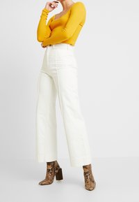 Vrouw in mosterdgele geribbelde trui, hoge taille brede witte broek en bruine slangenleer blokhakken enkel laarzen, staand.