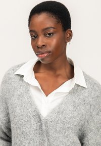 Cárdigan gris esponjoso con diseño de escote en V, usado sobre una camisa blanca con collar. Presenta un tejido texturizado y botones visibles.