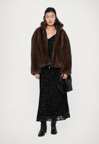 Cappotto di pelliccia marrone con colletto alto, indossato sopra un vestito di pizzo nero. L'outfit comprende una pochette nera e stivaletti, creando un look stratificato.