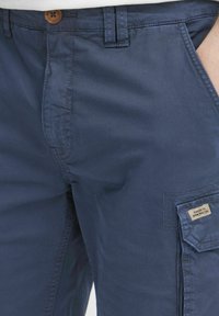 Sötétkék cargo nadrág sima pamutból, elülső zsebből és oldalsó felvarrt zsebből, amely címkét is tartalmaz. Gombos záródás a deréknál.