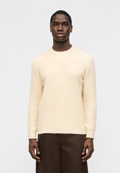 Man wearing a cream-colored ribbed sweater and dark brown pants, standing against a plain white background, facing forward.