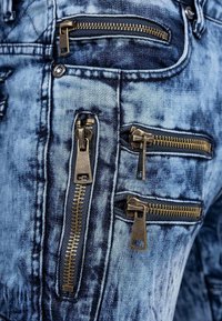 Close-up of blue denim jeans with five visible brass zippers on pocket areas.