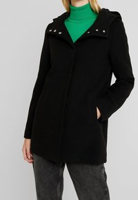 Černý kabát s kapucí, s stříbrnými druky u výstřihu a bočními kapsami, vyrobený z texturované látky. V kombinaci se zeleným rolákem.