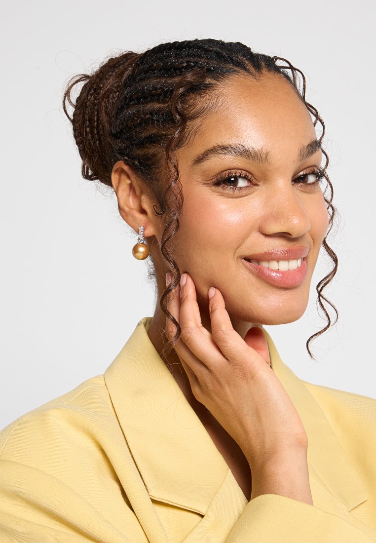 Pendientes de perlas doradas con un pequeño acento de cristal, llevados por una persona con un blazer amarillo, piel clara, cabello trenzado y rizos suaves.