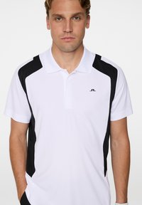 Vit pikétröja med svarta sidopaneler, korta ärmar och krage. Har en liten logotyp på bröstet. Slät textur.