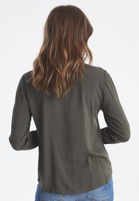Chemise à manches longues en vert olive foncé, fabriquée dans un tissu léger. Présente une texture lisse avec un ourlet arrondi et des manches à poignets.