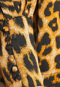 Section de blouse à motif léopard mettant en valeur un tissu orange et noir avec une manche froncée et cinq boutons le long du poignet.