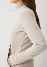 Giacca in fleece beige con colletto alto, tasche laterali, logo discreto sulla manica e texture liscia.