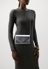 Srebrna metaliczna kopertówka z łańcuszkiem, charakteryzująca się eleganckim designem i ostrymi kątami, wyeksponowana na tle ciemnej prążkowanej tkaniny.