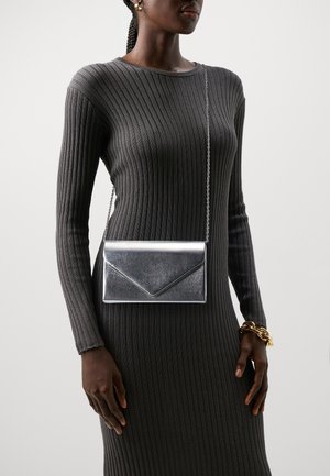 Srebrna metaliczna kopertówka z łańcuszkiem, charakteryzująca się eleganckim designem i ostrymi kątami, wyeksponowana na tle ciemnej prążkowanej tkaniny.