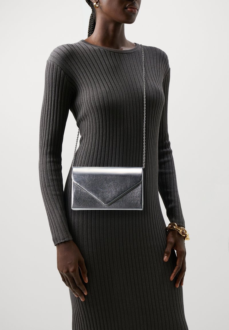 Srebrna metaliczna kopertówka z łańcuszkiem, charakteryzująca się eleganckim designem i ostrymi kątami, wyeksponowana na tle ciemnej prążkowanej tkaniny.