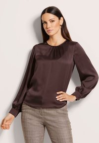 Frau mit langen dunklen Haaren, die eine dunkelbraune Satinbluse und karierte Hose trägt, posiert mit einer Hand auf der Hüfte vor einem hellen Hintergrund.